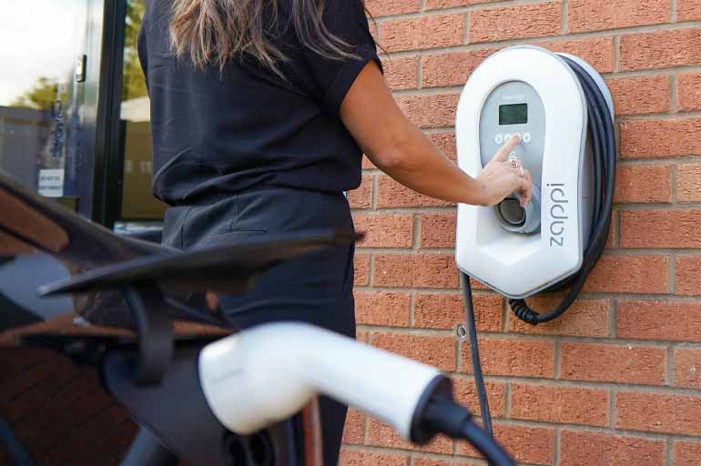 Eine Person drückt auf Tasten einer Wandladestation. Im Anschnitt ist ein ladendes Fahrzeug zu sehen.