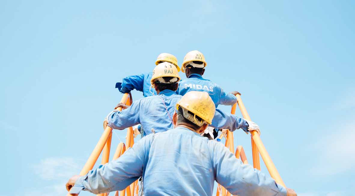 Vier Personen mit blauer Kleidung und gelbem Baustellenhelm steigen eine Leiter oder ein Gerüst auf einer Baustelle hoch