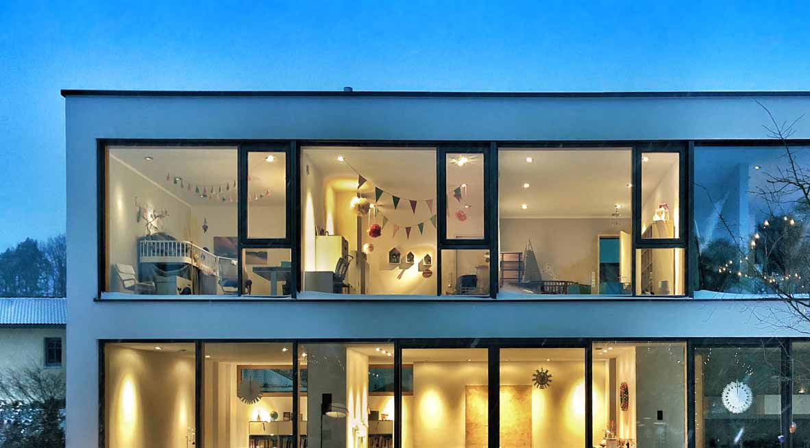 Großes Einfamilienhaus mit Glasfronten in der Abenddämmerung. Es brennt Licht im ganzen Haus.