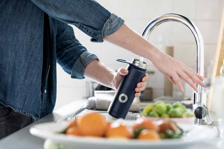 Hände einer Frau halten eine Trinkflasche, die an einem Spülbecken in einer Küche mit Wasser aus dem Wasserhahn gefüllt wird.