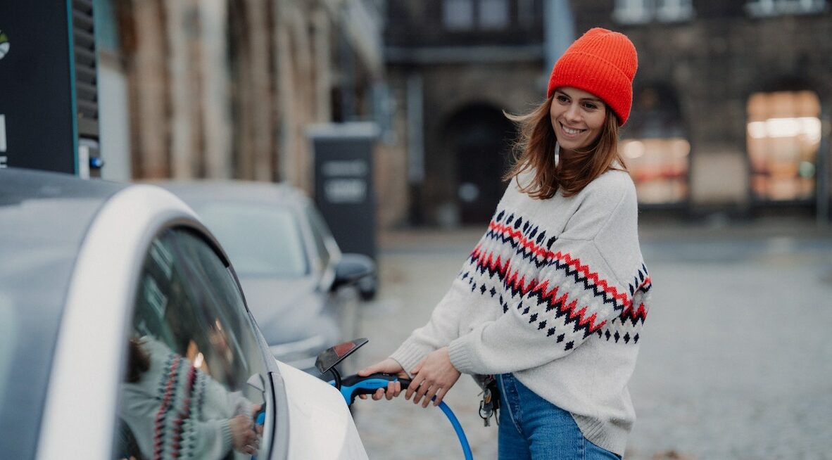 Eine Frau mit roter Mütze und Wollpulli steckt das Ladekabel in ihr Elektrofahrzeug
