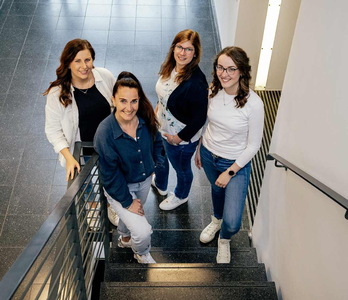 Teambild von 4 Mitarbeiterinnen aus dem Kundenzentrum der StadtWerke Rösrath. Sie stehen am Fuße der Treppe im Eingangsbereich und lachen sehr freundlich und sympathisch in die Kamera.