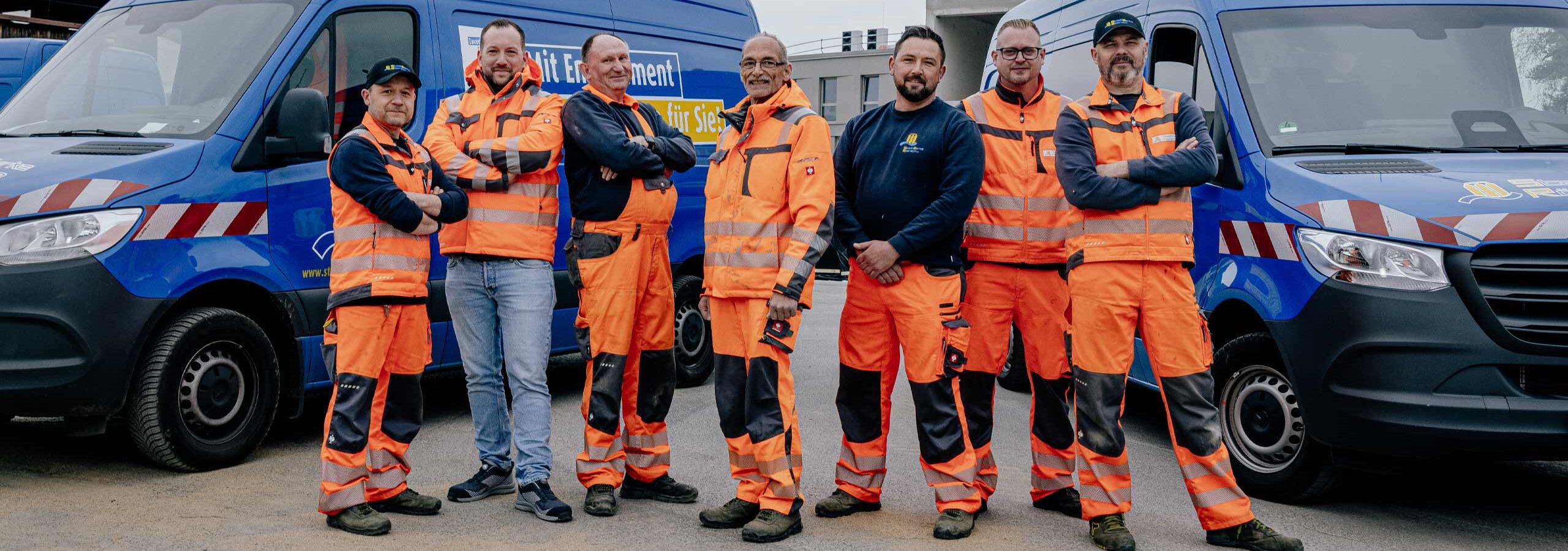 Gruppenbild von 7 Mitarbeitern aus der Abteilung Trinkwassernetz der StadtWerke Rösrath. Sie stehen von ihren Fahrzeugen und tragen orangefarbene Arbeitskleidung.